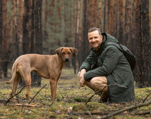 Ein Rhodesian Ridgeback mit seinem Besitzer im herbstlichen Wald, Empfehlung für Tierhalter-Haftpflichtversicherung und OP-Versicherung im Vergleich zur Voll-Krankenversicherung.